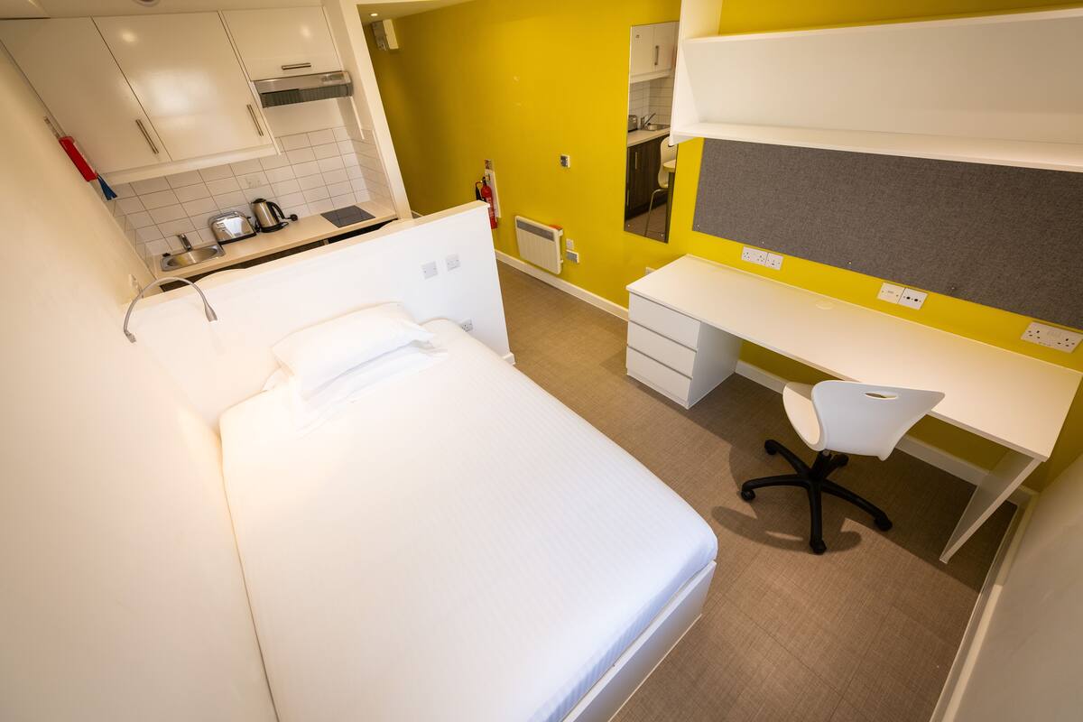 Quarto de hotel com cama, escrivaninha, cadeira, cozinha compacta e banheiro.
