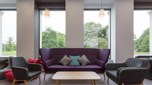 A modern living room with a purple sofa, two armchairs, and a wooden coffee table. Large windows provide a view of greenery outside. - University of Bath City Accommodation (Bath)