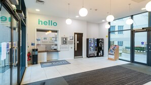 A reception area with a 'hello' sign, a reception desk, a vending machine, a magazine rack, and a large window offering a view outside. - University of Bath City Accommodation (Bath)