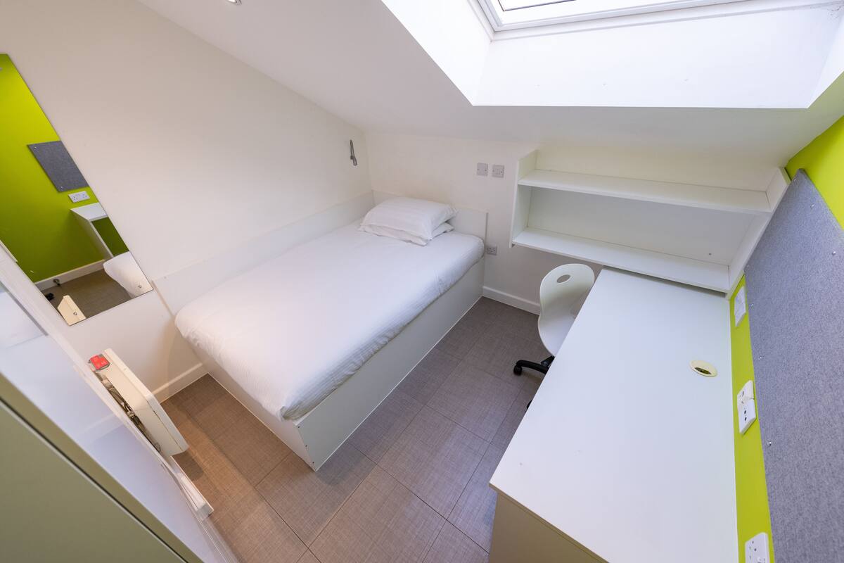 Quarto pequeno com cama, escrivaninha e banheiro.