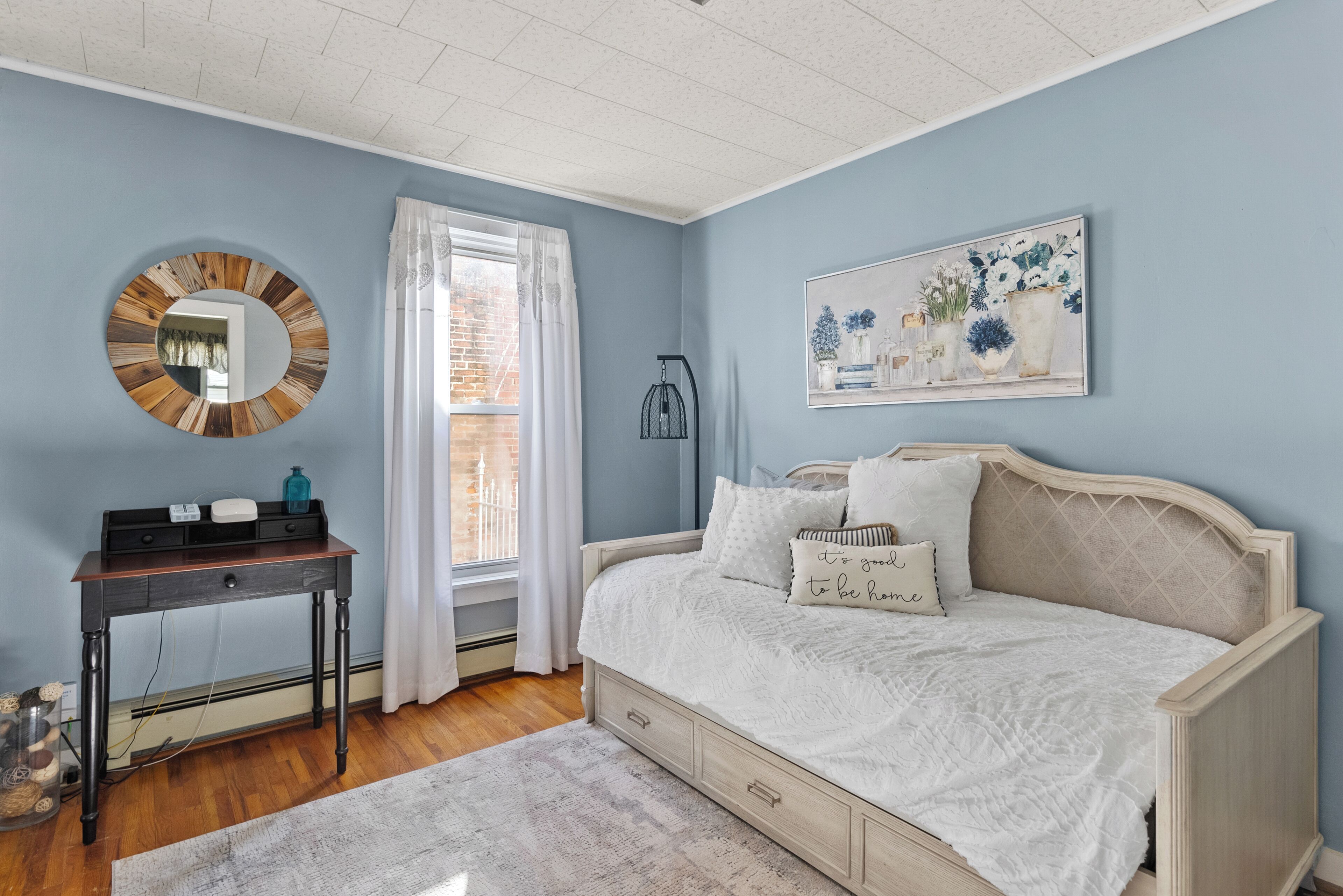 A bedroom with a bed, a desk, a mirror, and a window with curtains.