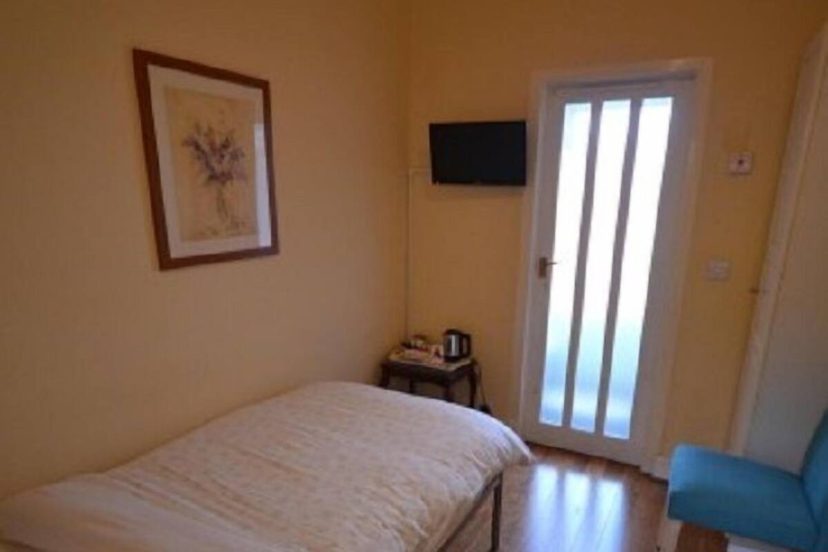 A bedroom with a bed, a chair, a small table with a kettle, a framed picture on the wall, and a door leading to a balcony.