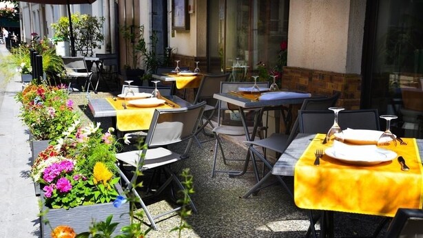 Un área de comedor al aire libre con mesas dispuestas para dos, cubiertas con manteles amarillos, rodeada de plantas y flores en macetas.