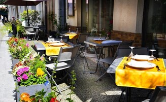 An outdoor dining area with tables set for two, covered with yellow tablecloths, and surrounded by potted plants and flowers.