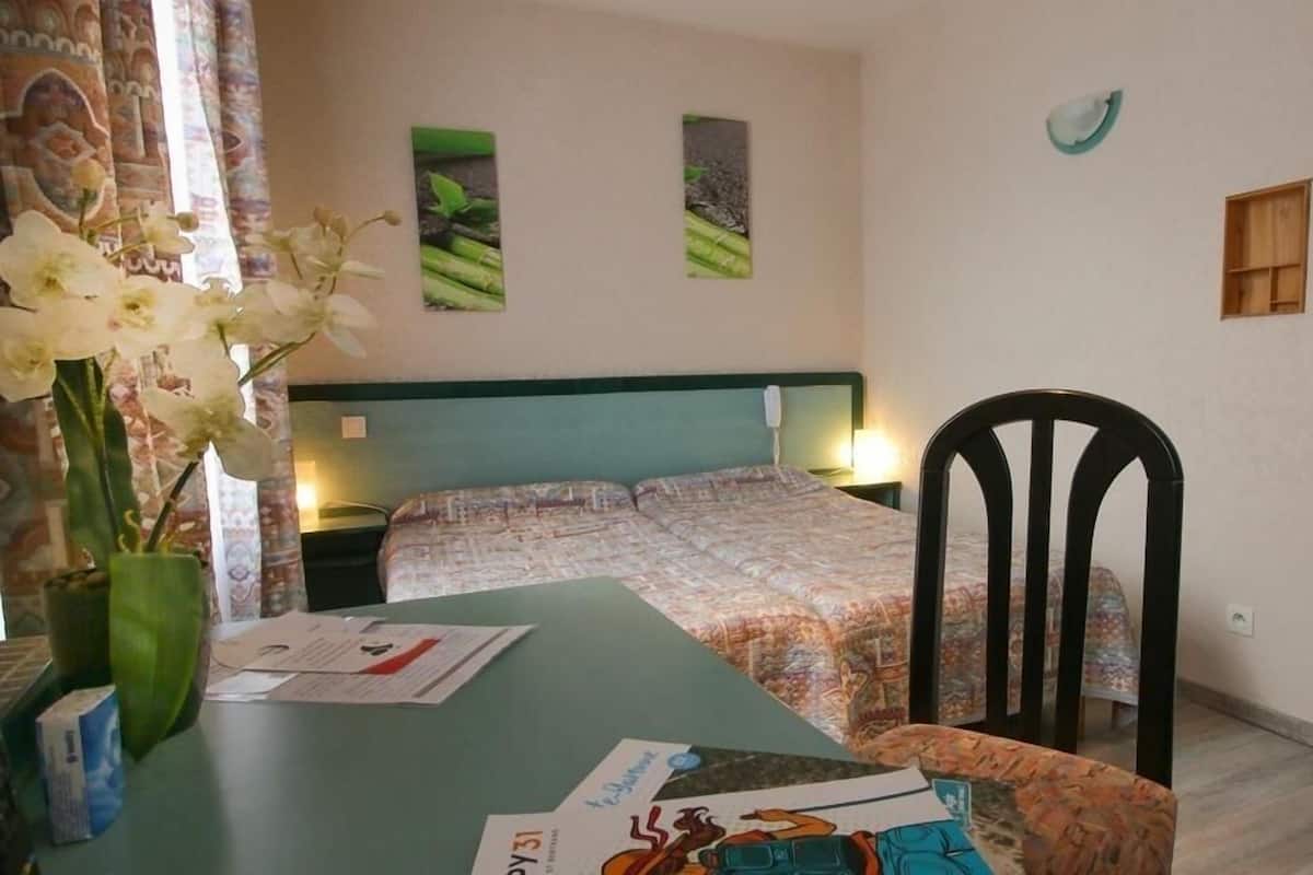 A bedroom with a bed, a chair, a table with papers, and a vase with flowers.