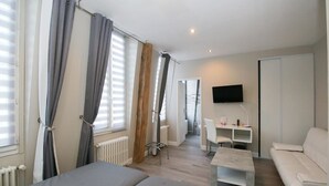 Habitación moderna y luminosa con cama, sofá blanco, una mesita y televisor montado en la pared.