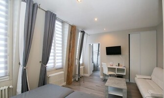 A modern, well-lit room with a bed, a white sofa, a small table, and a TV mounted on the wall.