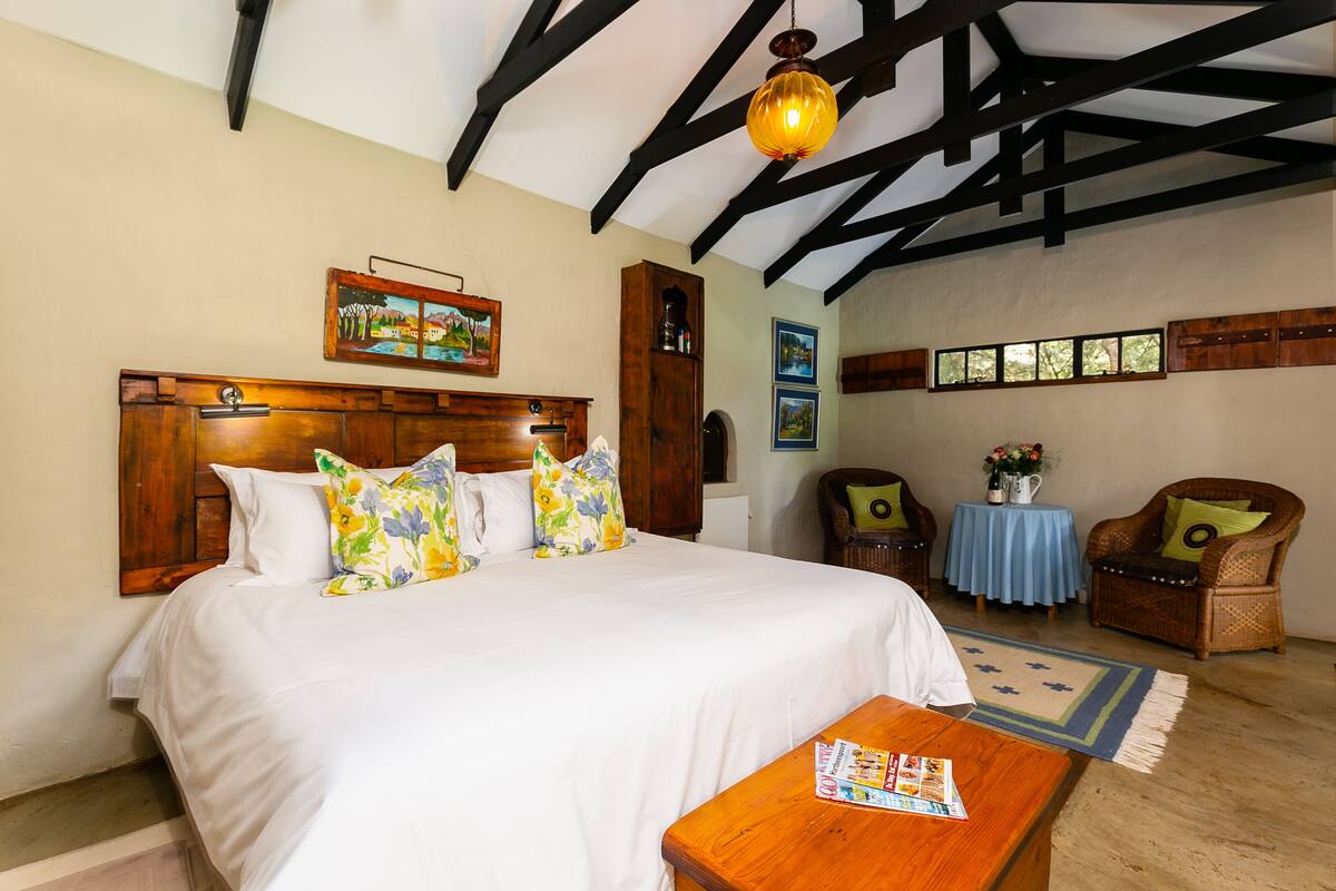 Un dormitorio con una cama grande, cabecera de madera, una mesa de madera con una revista, mantel azul, sillas de mimbre y un cuadro en la pared.