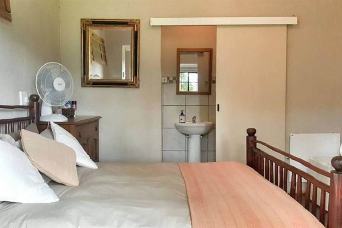 Un dormitorio con cama, mesita de noche, espejo, ventilador y vista a un baño con lavamanos y espejo.