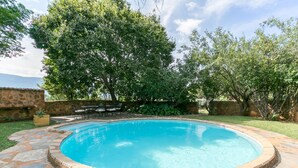 A swimming pool with a stone border, surrounded by a stone wall and a grassy area with trees. - La Bastide Guesthouse (Hartbeespoort)