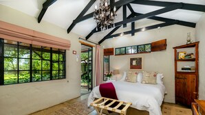 A bedroom with a large bed, a wooden bench, a chandelier, and a view of greenery through the window. - La Bastide Guesthouse (Hartbeespoort)