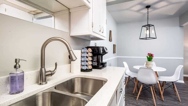 Una cocina moderna con un lavamanos de acero inoxidable, una cafetera y un comedor con sillas blancas y una pequeña mesa.