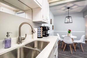 A modern kitchen with a stainless steel sink, a coffee machine, and a dining area with white chairs and a small table.