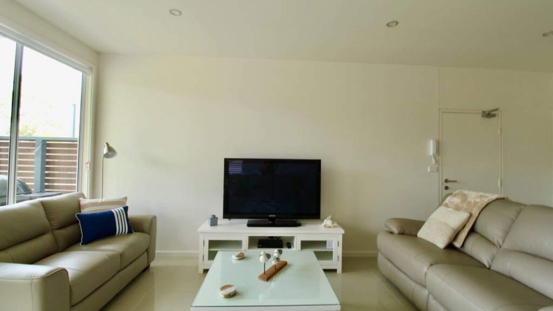 Uma sala de estar com uma TV sobre um suporte branco, dois sofás bege, uma mesa de centro com objetos decorativos e uma janela grande.