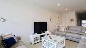 A living room with a flat-screen TV on a white stand, a grey sofa, a white coffee table, and a lamp. - Avalon Apartment by Book Jervis Bay (Huskisson)