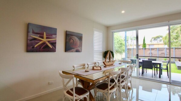 Un comedor con un ventanal que da a un patio exterior. Hay dos cuadros en la pared, una estrella de mar y una concha.