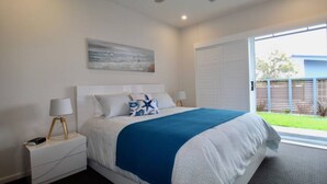 A bedroom with a large bed, two bedside tables with lamps, a window with a view of the outside, and a painting on the wall. - Avalon Apartment by Book Jervis Bay (Huskisson)