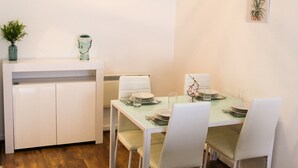 Ein Essbereich mit einem weißen Tisch, gedeckt für vier Personen, weißen Stühlen, einem weißen Schrank und einer kleinen Pflanze auf dem Schrank.