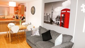 A living room with a grey sofa, white chairs, a wooden dining table, and a kitchen in the background.