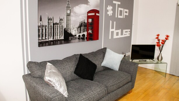 A grey sofa with black and white cushions, a flat-screen TV on a glass table, and a wall with a cityscape print and the hashtag #TopHouses.