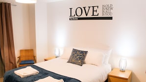 A neatly made bed with a dark blue bedspread and white pillows, a wall with a quote from The Beatles, a chair with a blue cushion, and bedside lamps on wooden nightstands.