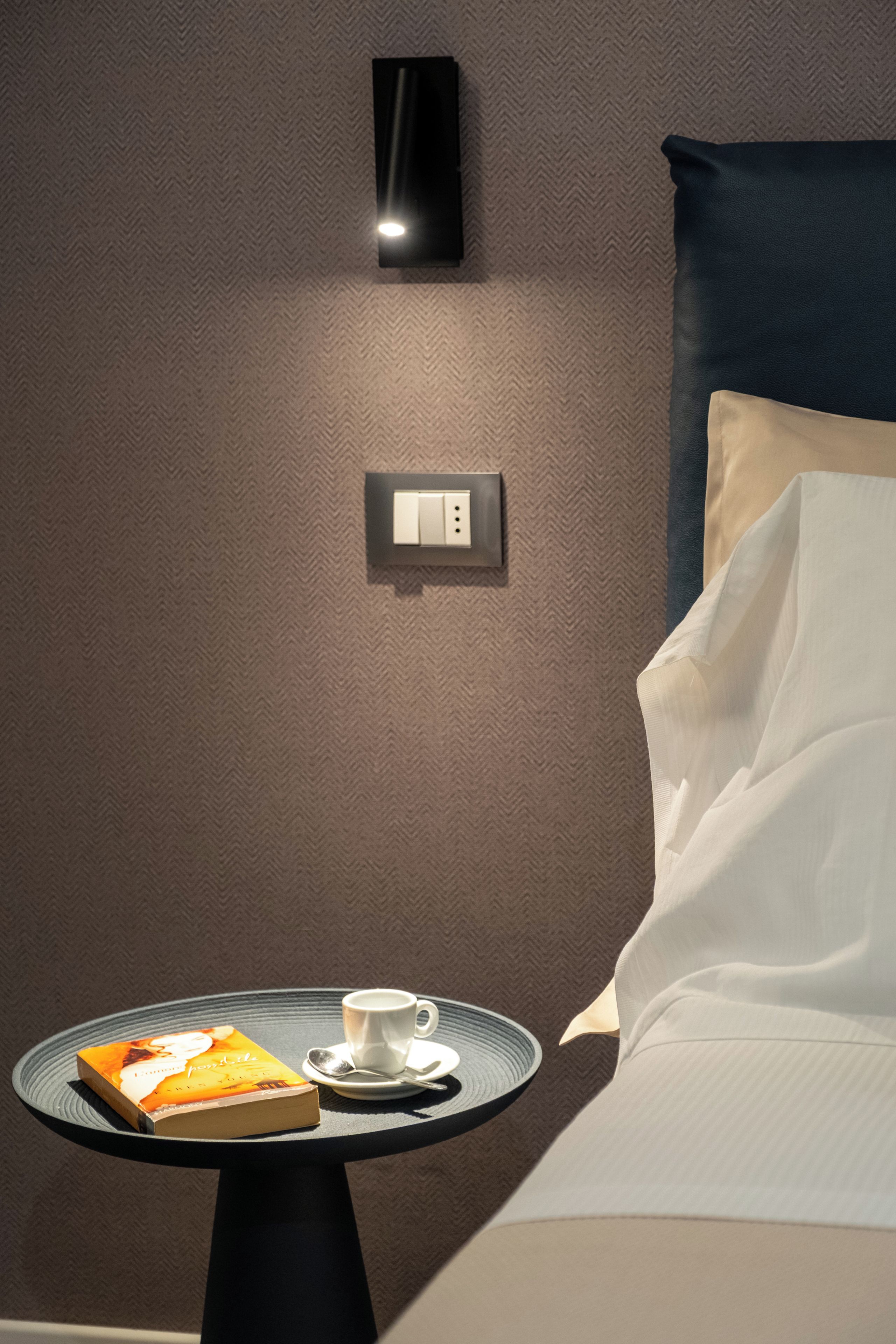 Een hotelkamer met een bed, een nachtkastje met een boek en een kopje, een wandlamp en een stopcontact.