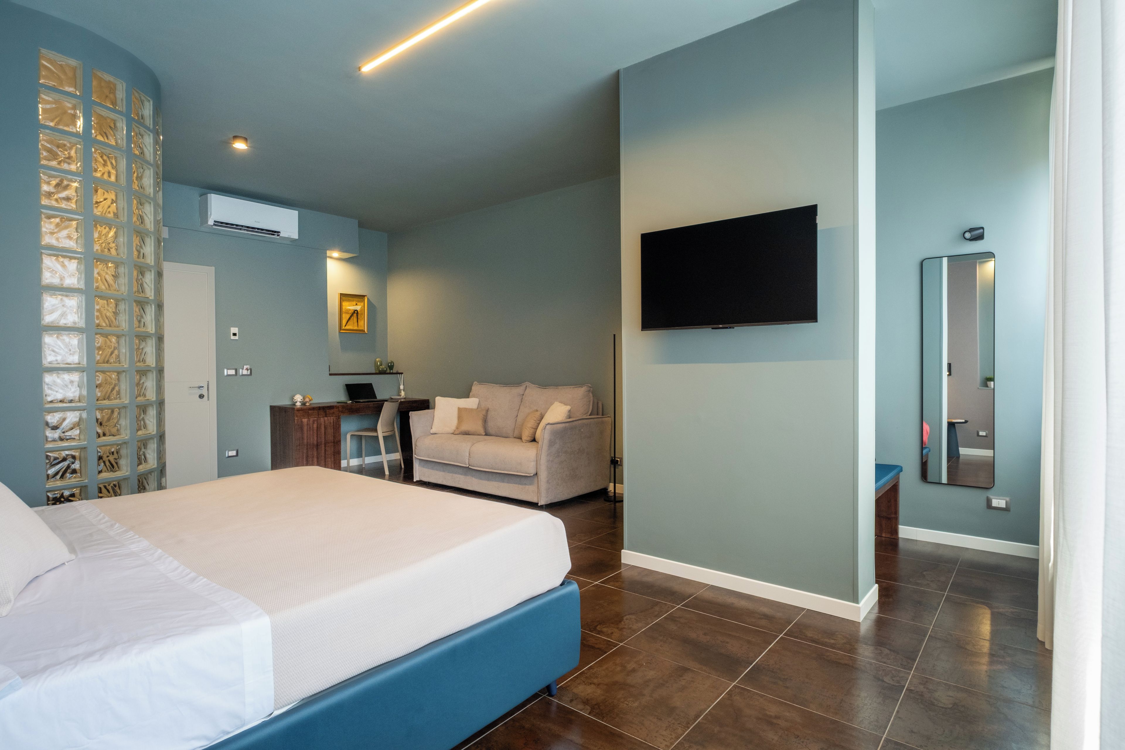 A modern bedroom with a bed, a desk, a sofa, a TV, and a bathroom mirror.