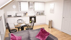 Une cuisine compacte avec des meubles blancs, un four à micro-ondes et un grille-pain. On y trouve une table à manger avec des chaises, ainsi qu’un canapé gris agrémenté d’un plaid violet.