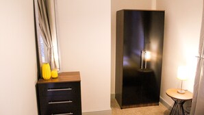 A hotel room with a dark wardrobe, a mirror with curtains, a small table with a lamp, and a yellow vase on the dresser. - Mersey Place (Liverpool)