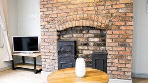 A room with a brick fireplace, a television on a stand, and a round table with a white vase. - Mersey Place (Liverpool)