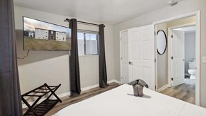A bedroom with a bed, a TV mounted on the wall, a door leading to a bathroom, and a window with curtains. - Lansing Layover #4 | New York State of Mind (Lansing)