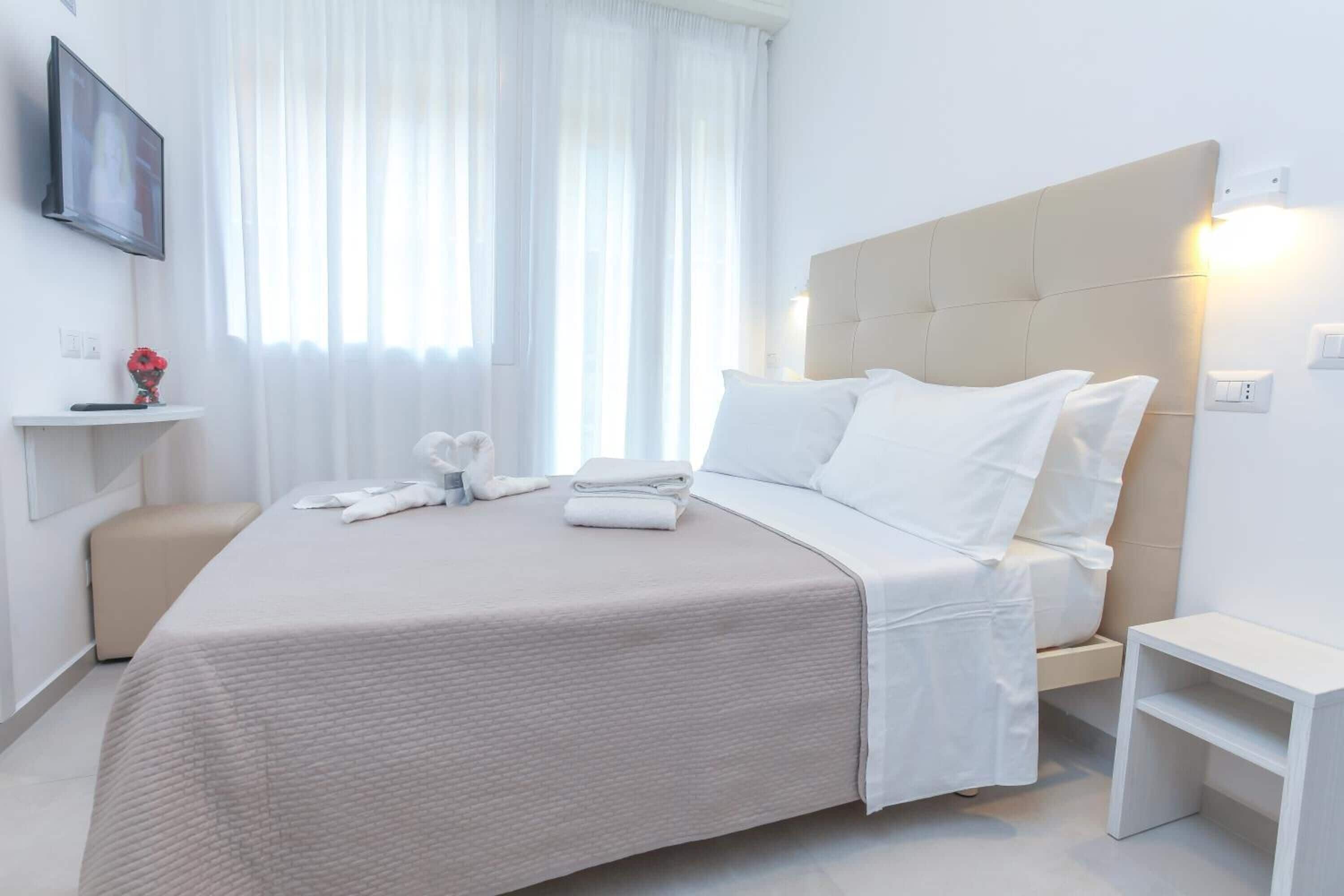 A modern hotel room with a large bed, a bedside table, a TV mounted on the wall, and a window with sheer curtains.