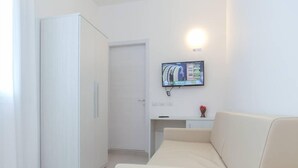 A modern, minimalist room with a white wardrobe, a beige sofa, a small table, and a mounted TV. - Hotel Tourist Meuble (Rimini)