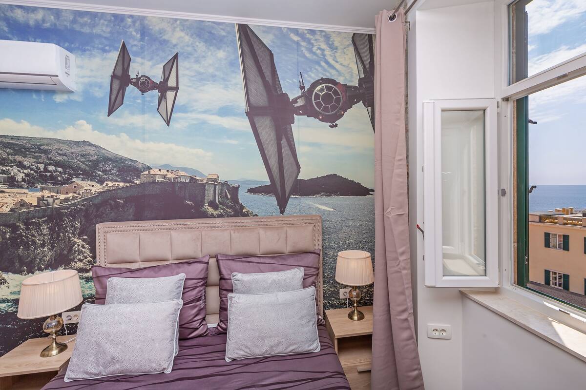 Une chambre à coucher avec un lit, des oreillers, des lampes de chevet, une fenêtre donnant sur la mer et les bâtiments, et une fresque murale représentant un paysage avec des objets volants.