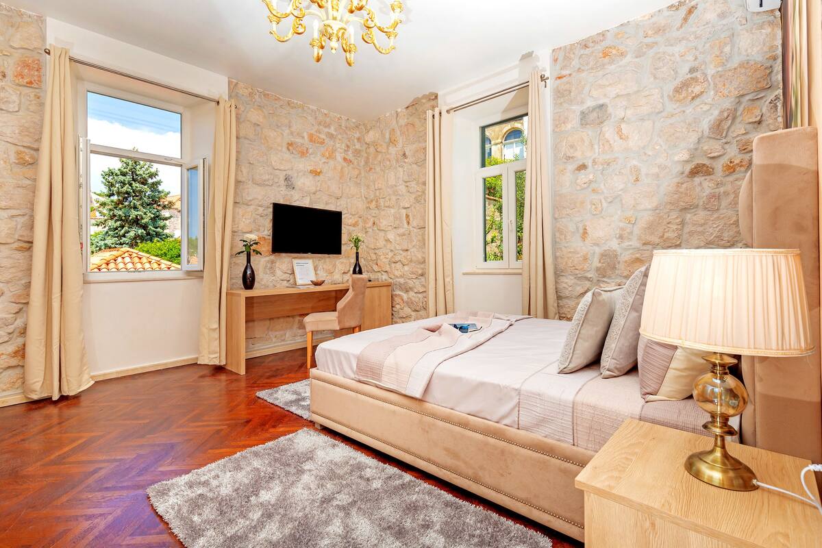 Une chambre avec un mur en pierre, un lit, un bureau avec un ordinateur et une fenêtre avec des rideaux.