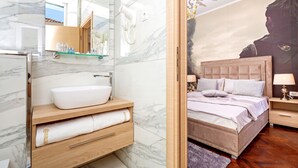 Een moderne badkamer met een marmeren wastafel, een houten opbergmeubel en een grote spiegel. De slaapkamer heeft een groot bed met hoofdbord, een nachtkastje en een muurschildering.