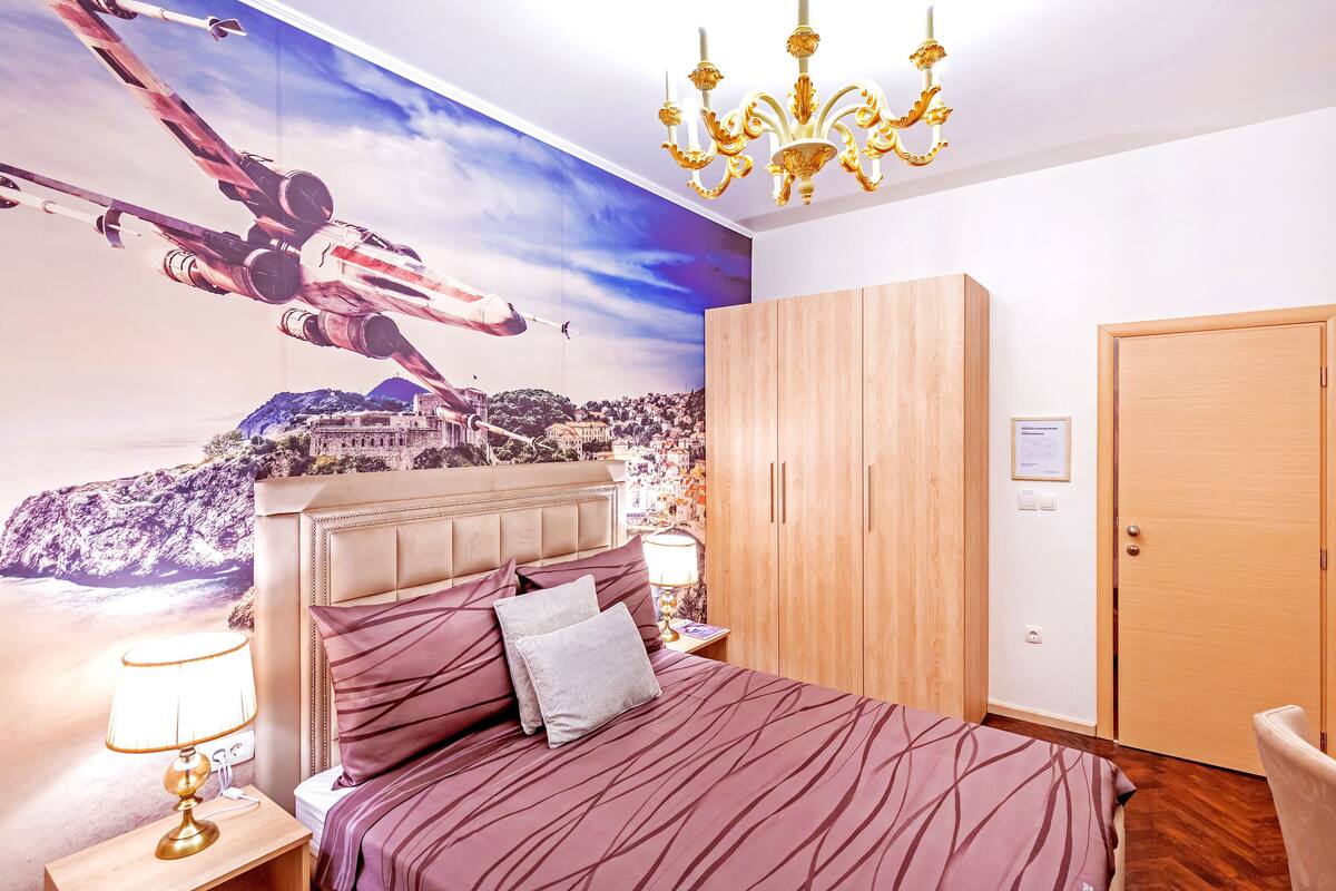 Une chambre à coucher avec un grand lit, une table de chevet, un lustre, une armoire et une fresque murale représentant un paysage urbain.