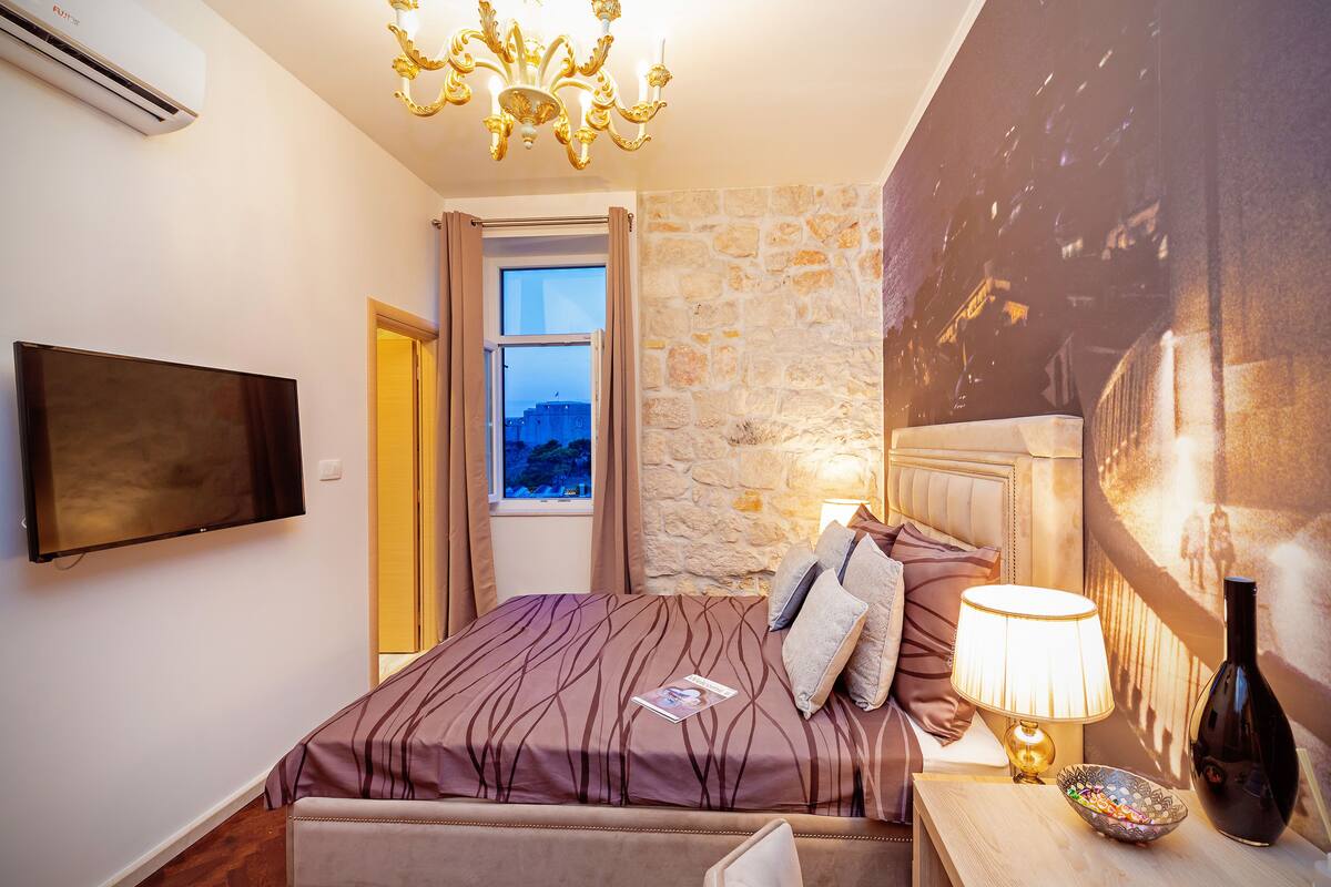 Une chambre à coucher avec un grand lit, un téléviseur à écran plat, un lustre, un mur en pierre et une fenêtre avec des rideaux.