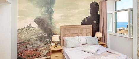 Een slaapkamer met een groot muurschildering van een stadsgezicht en een figuur, een bed met een beige hoofdbord, twee nachtkastjes met lampen, en uitzicht op de zee door een raam.