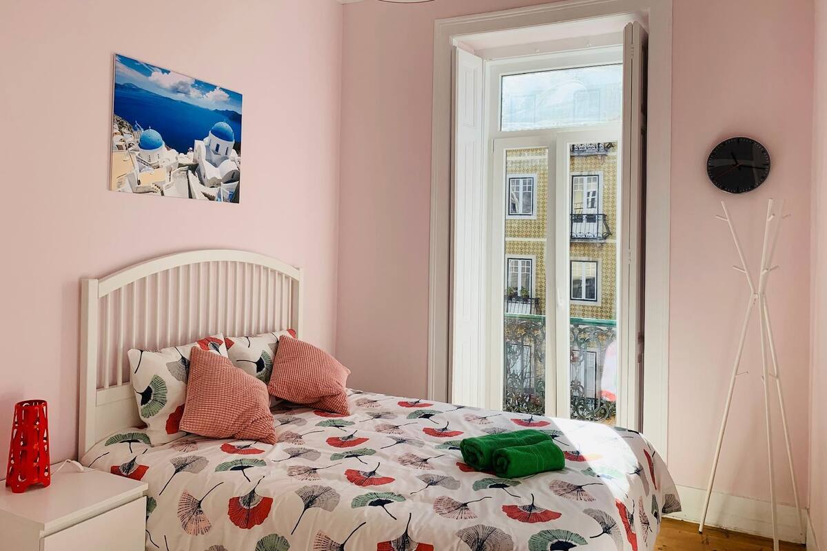 A bedroom with a bed, a nightstand, a red vase, a painting, a window, and a clock.