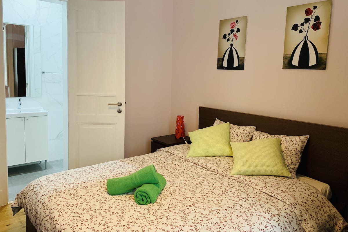 A bedroom with a bed, a nightstand, a red vase, and two wall art pieces.