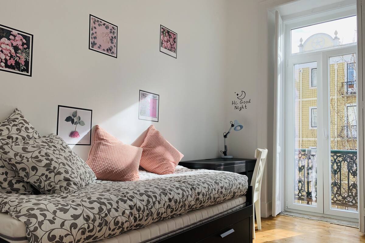 A bedroom with a bed, a desk, and a chair. There are pictures on the wall and a window with a view of buildings.
