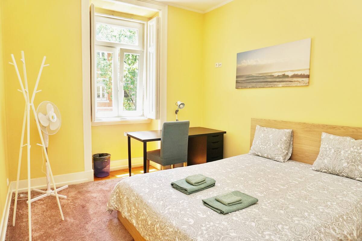 A bedroom with a bed, a desk, a chair, a standing fan, and a window with a view of greenery.