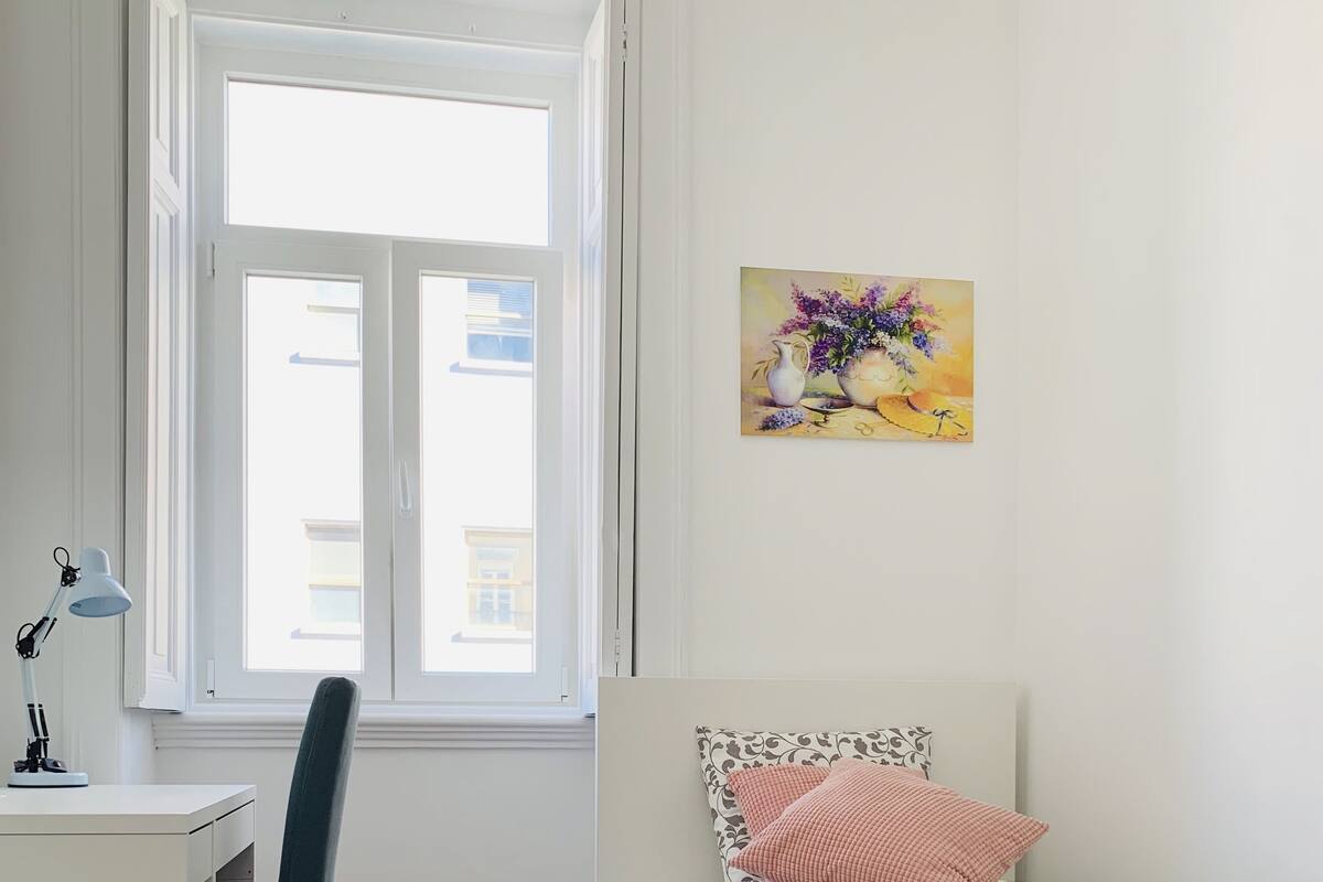 A bedroom with a bed, a desk, a chair, a window, and a painting on the wall.