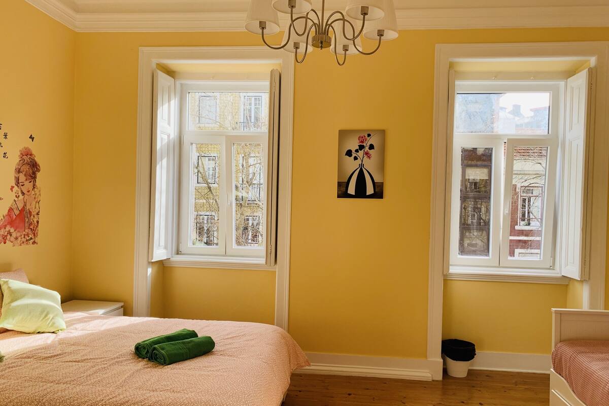 A bedroom with two windows, a bed with a pink bedspread, a green towel, and a painting on the wall.