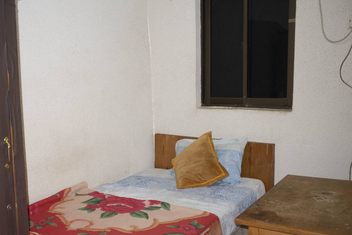A small, simple room with a bed, a wooden nightstand, and a chair.