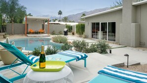 Un área de piscina moderna al aire libre con sillones de descanso, una mesa con una botella y vasos, y un espacio de estar cubierto.