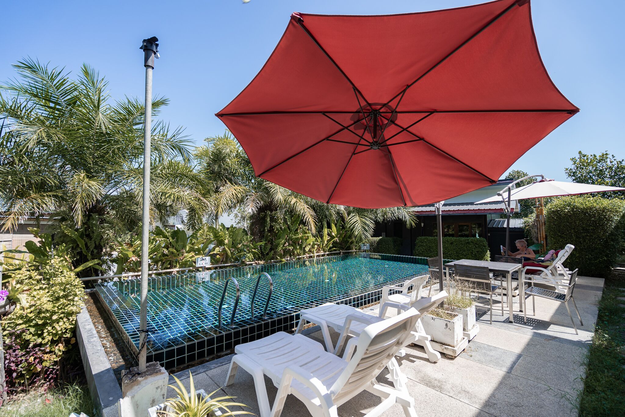 Une piscine avec un parasol rouge, des chaises longues blanches et une clôture en métal.