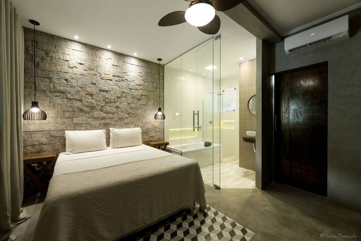 A modern bedroom with a stone wall, a large bed, a glass shower enclosure, and a ceiling fan.