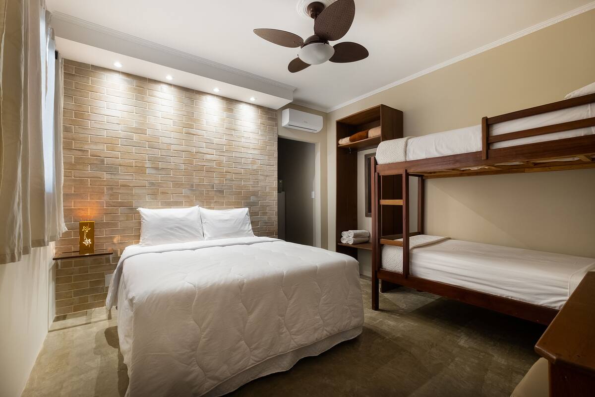 A hotel room with a brick-patterned wall, a bed with white bedding, a ceiling fan, and a bunk bed.